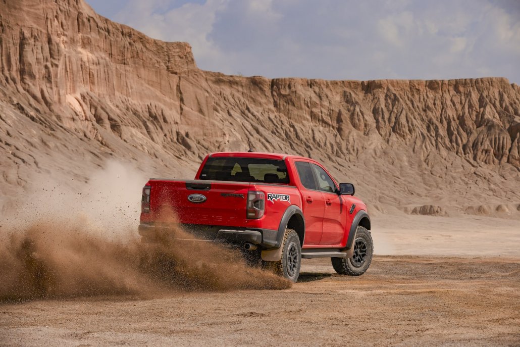 Ein orangener Ford Ranger Raptor 2022 driftet durch eine Wüste. Im Hintergrund befindet sich eine große, rote Felsfront unter dunstigem Himmel.