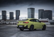 Ein gelber BMW X4 M Competition steht auf einer Asphaltfläche vor Hochhäusern.