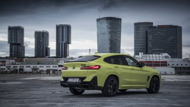 Ein gelber BMW X4 M Competition steht auf einer Asphaltfläche vor Hochhäusern.