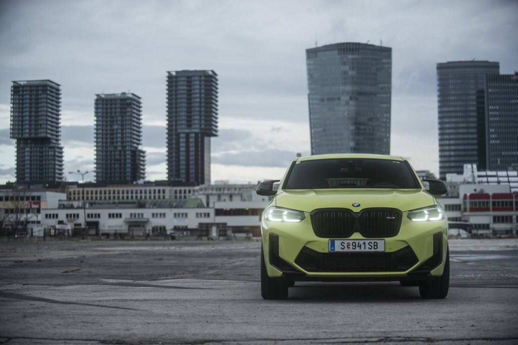Ein gelber BMW X4 M Competition steht auf einer Asphaltfläche vor Hochhäusern.