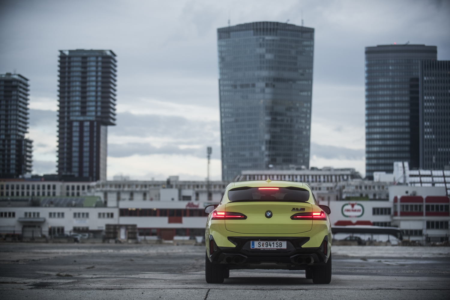 Ein gelber BMW X4 M Competition steht auf einer Asphaltfläche vor Hochhäusern.