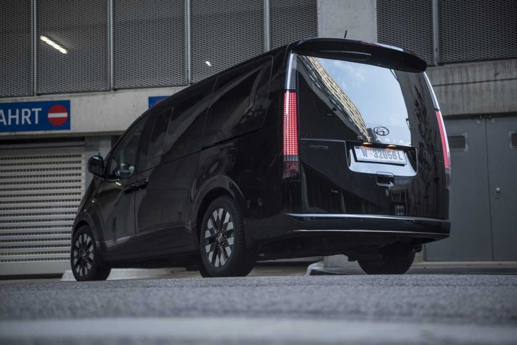 Ein schwarzer Hyundai Staria mit eingeschalteten Scheinwerfern steht vor einer Garagenein- und -ausfahrt.