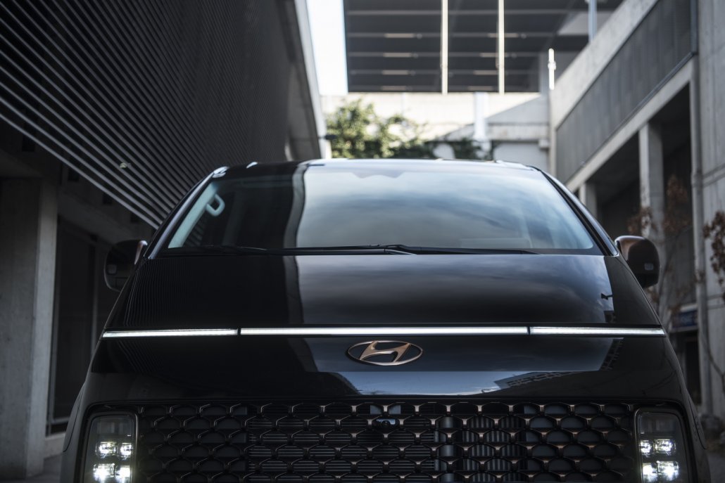 Die Front eines stehenden, schwarzen Hyundai Staria mit eingeschalteten Scheinwerfern, bronzenen Akzenten an Logo und Kühlergrill und ein horizontal durchgehendes Leuchtenband.
