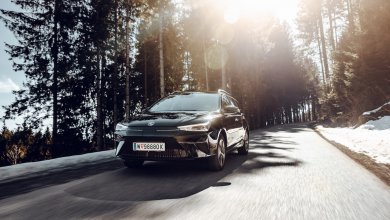 Ein schwarzer MG5 Electric fährt an einem sonnigen Tag auf einer Landstraße im Wald zwischen Bäumen. Neben der Landstraße liegt etwas Schnee.