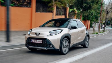 Ein champagnerfarben-schwarzer Toyota Aygo X fährt auf einer urbanen Straße einer orangenen Mauer entlang.