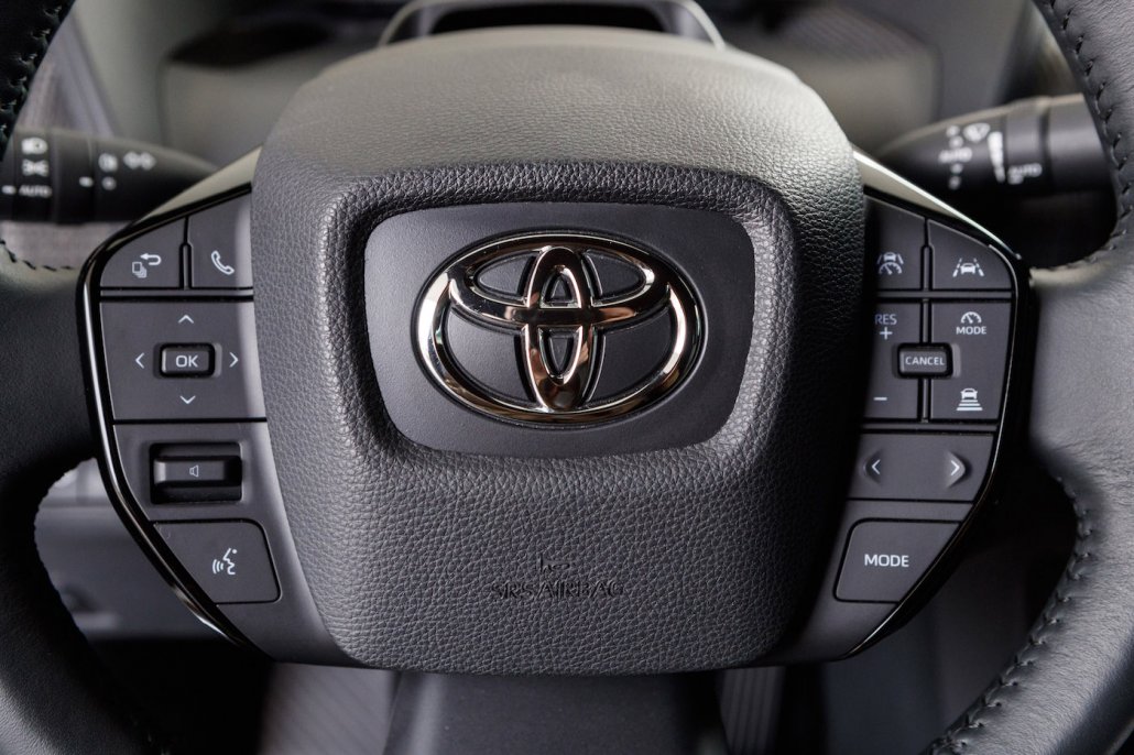 Detailaufnahme eines schwarzen Toyota bZ4X-Lenkrads. Links und rechts vom Lenkradtopf befinden sich viele Knöpfe für Assistenzsysteme, Telefonie und zum Bedienen der digitalen Armatur.