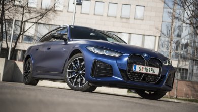 Ein blauer BMW i4 M50 steht auf einem Parkplatz vor einem grauen Gebäude und kahlen Bäumen.