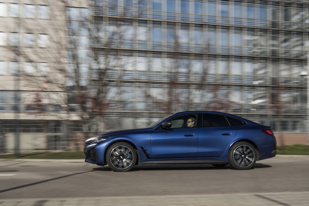Ein blauer BMW i4 M50 fährt auf einem Parkplatz vor kahlen Bäumen und einem gläsernen Gebäude.