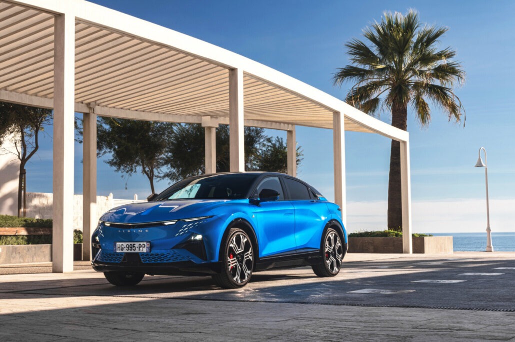 Ein blauer Alpine-SUV in Dreiviertelansicht vorne links steht unter einer modernen Pergola an einer Promenade mit Meerblick und Palme.