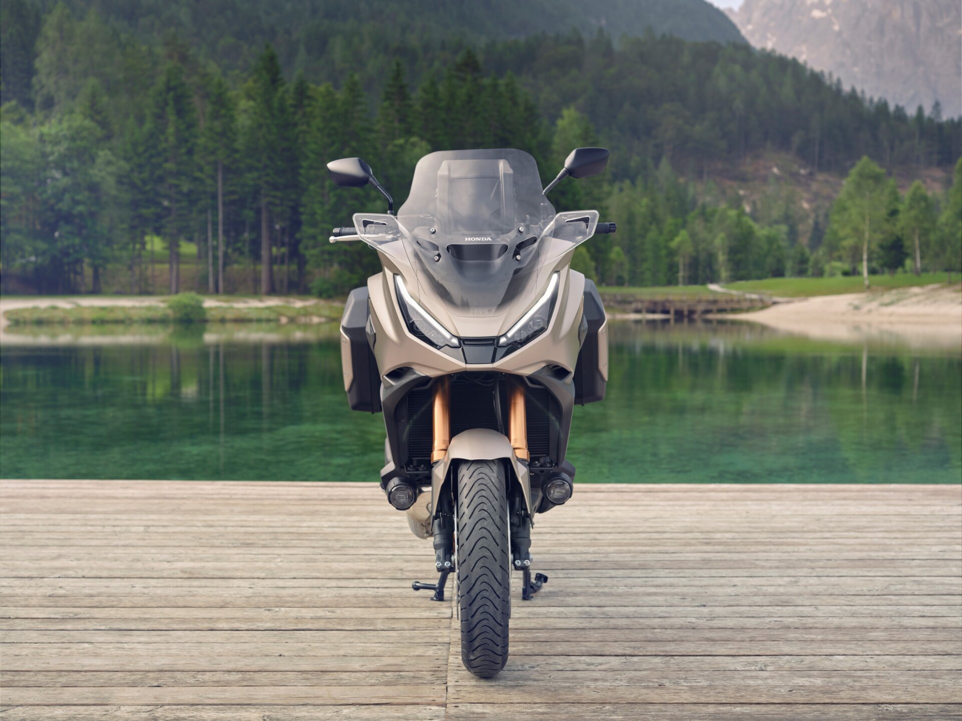Ein beiges Honda-Motorrad in Frontalansicht steht auf einem Holzsteg vor einem See und bewaldeten Bergen.