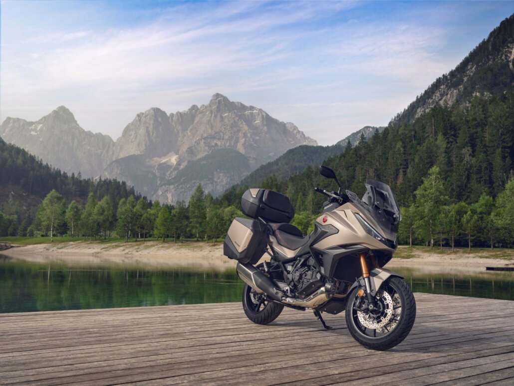 Ein goldfarbenes Motorrad in Dreiviertelansicht vorne rechts auf einem Holzsteg vor einer Berg- und See-Landschaft.