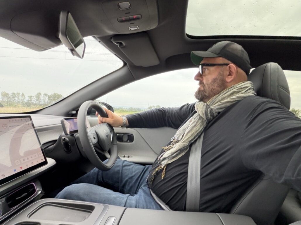 Der Innenraum eines Autos zeigt einen Fahrer auf dem Vordersitz in Fahrtrichtung, aufgenommen aus der Innenperspektive mit Blick auf das Armaturenbrett, das Lenkrad und den Touchscreen.