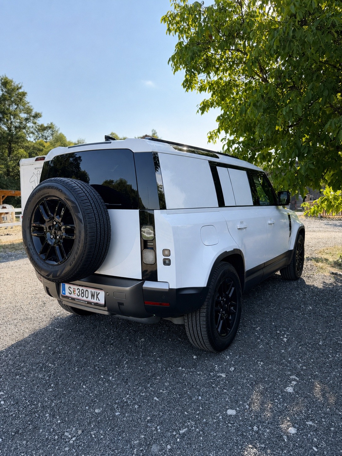 Weißer Geländewagen mit Ersatzrad am Heck auf einem Schotterplatz.
