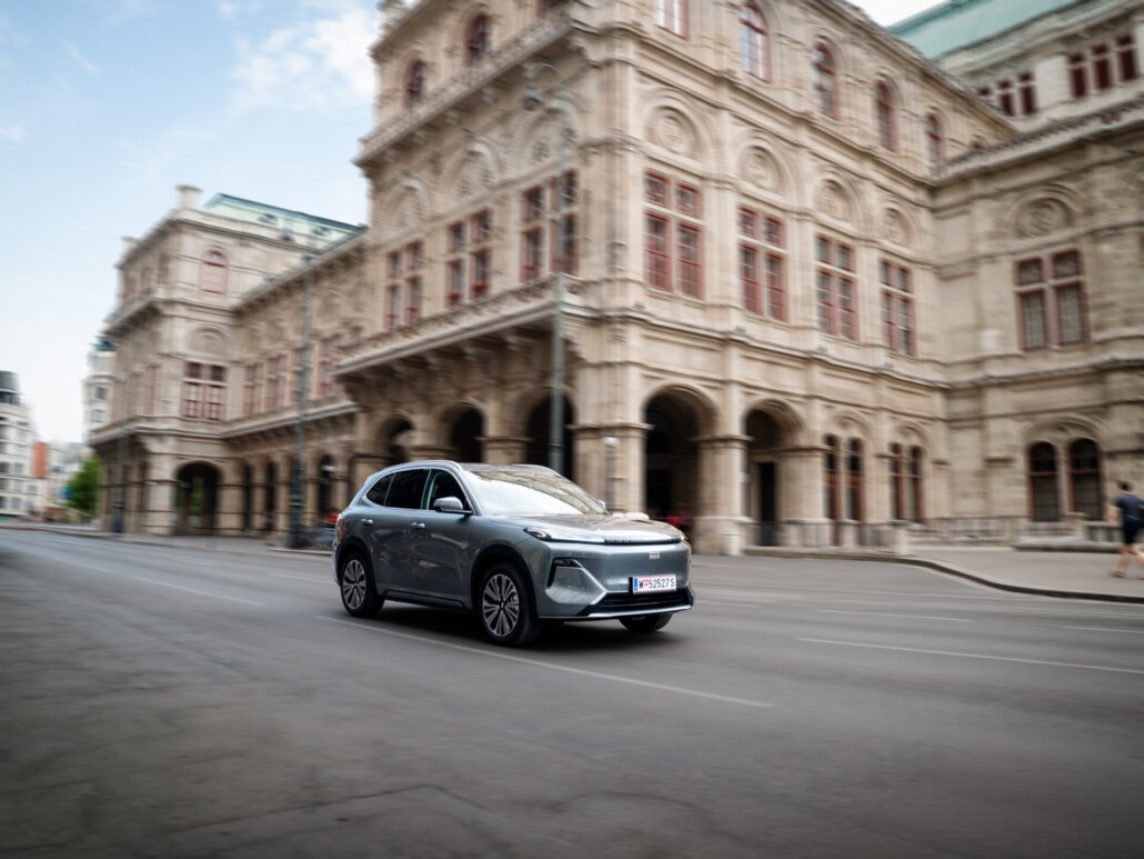 Ein grauer Kompakt-SUV in Dreiviertelansicht vorne rechts fährt auf einer Straße vor einem großen historischen Gebäude mit Arkaden.