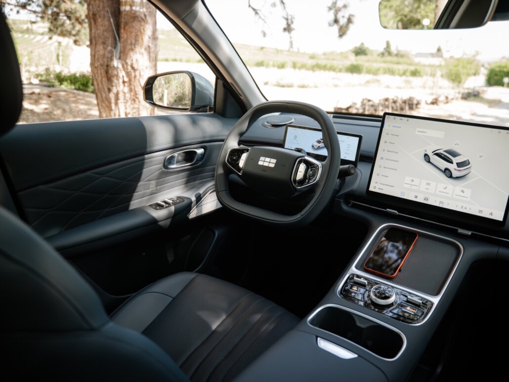 Der dunkelgraue Innenraum eines Autos zeigt das Armaturenbrett mit Lenkrad, Infotainment-Bildschirmen und Mittelkonsole aus der Perspektive vom Fahrersitz, im Hintergrund ist eine Landschaft mit Bäumen erkennbar.