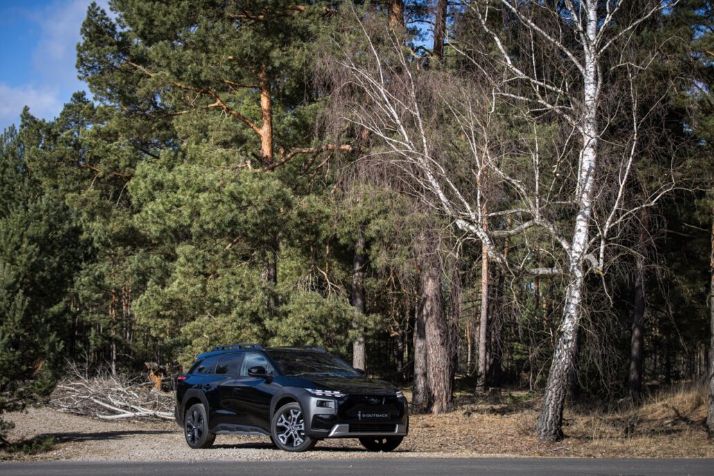 Zu sehen ist ein schwarzer SUV mit dem Kennzeichen-Schriftzug „E-OUTBACK“ in Dreiviertelansicht von vorne links, abgestellt auf einem Schotterstreifen vor einer Waldkulisse.