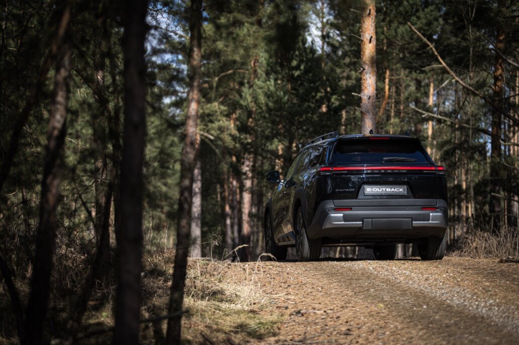 In der Heckansicht mit sichtbarer linker Flanke steht ein schwarzes SUV mit dem Schriftzug „E-OUTBACK“ auf einem unbefestigten Waldweg.