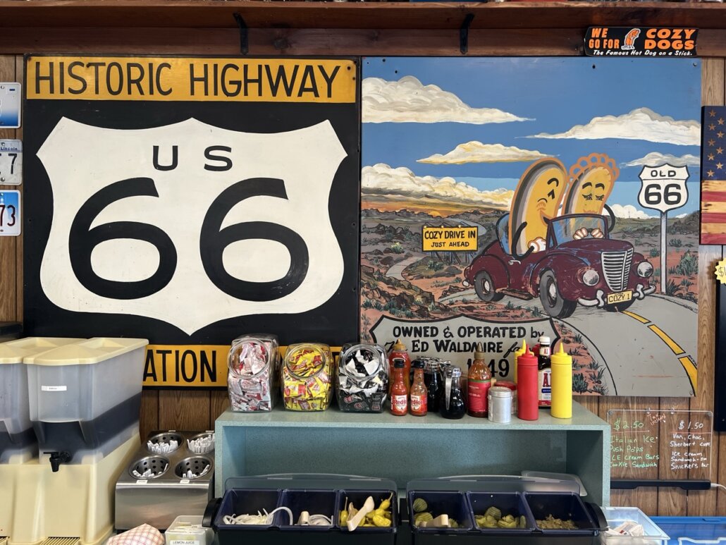 Eine gemalte Illustration zeigt ein rotes Oldtimer-Cabriolet in einer Landschaft aus der Dreiviertelansicht vorne rechts auf einem Plakat neben einem US-Route-66-Schild.