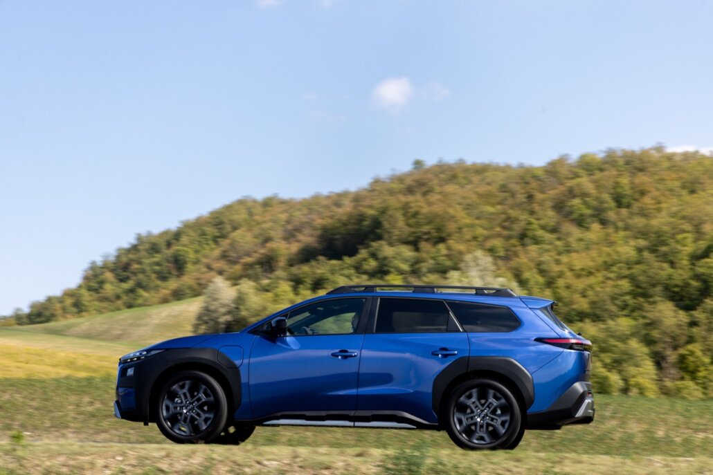 Ein blauer SUV in Seitenansicht fährt auf einer Landstraße vor einer bewaldeten Hügellandschaft.