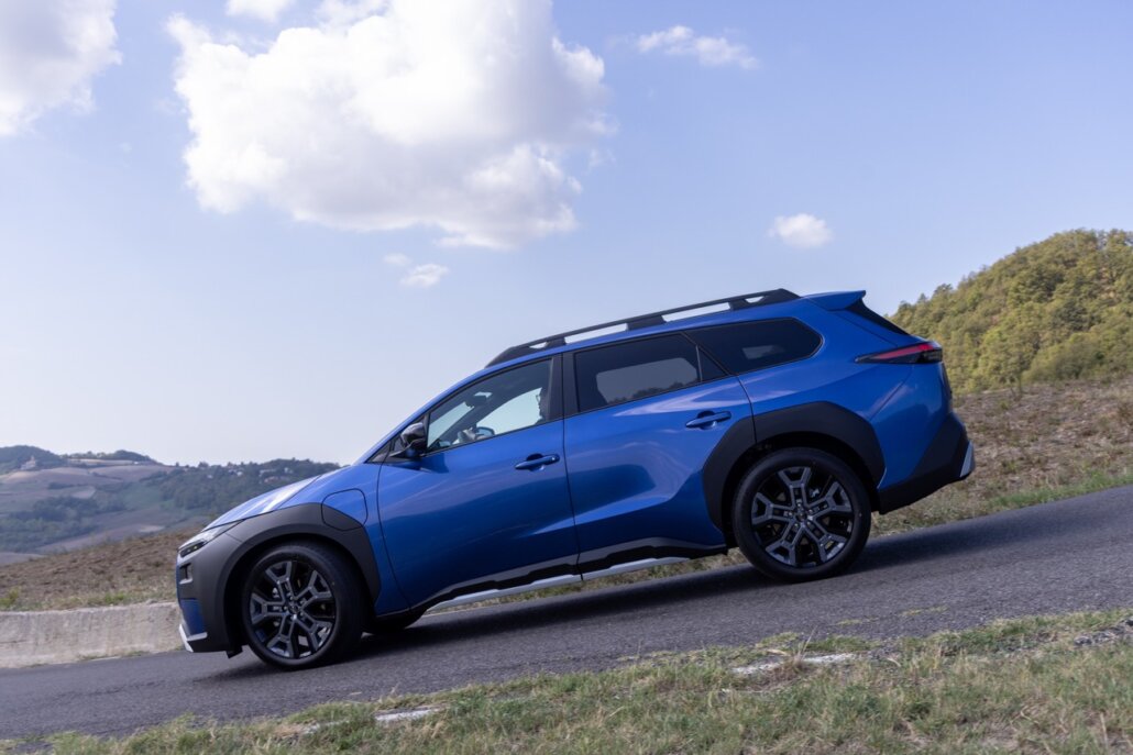 Ein blauer SUV in Seitenansicht steht auf einer Asphaltstraße in einer hügeligen Landschaft unter blauem Himmel.