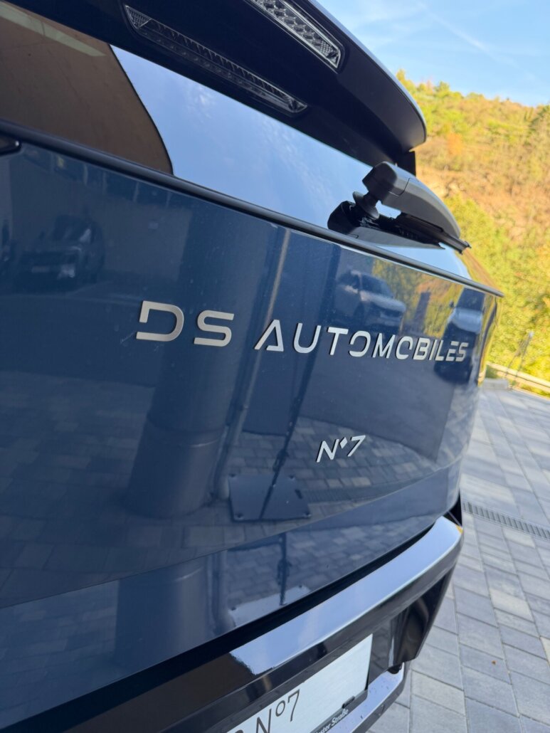 Ein Detail des Hecks eines dunkelblauen DS Automobiles N°7 SUV in Dreiviertelansicht, aufgenommen im Freien auf einem gepflasterten Platz vor einer Grünanlage.