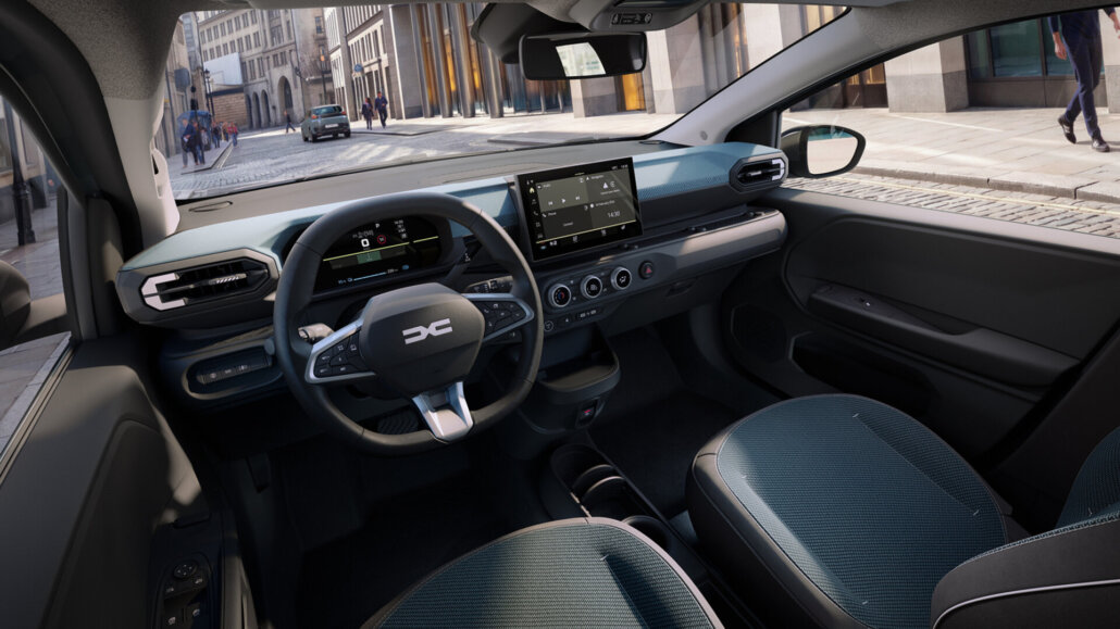 Der Innenraum eines dunkelgrauen Dacia-Kleinwagens mit blau abgesetzten Sitzen und Armaturenbrett, fotografiert aus der Perspektive des Beifahrers mit Blick auf Cockpit und Straße.