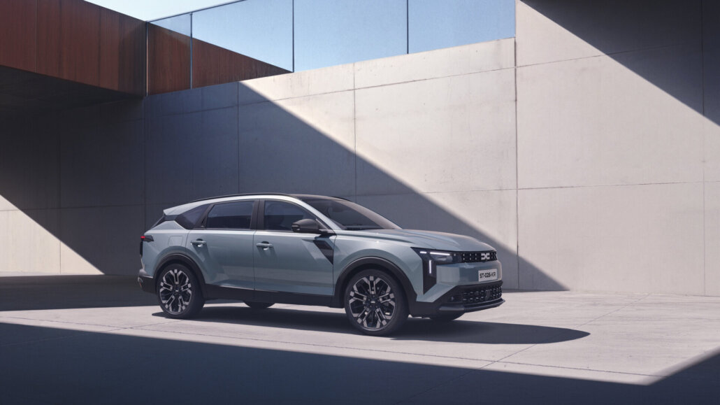 Ein hellblauer Dacia SUV in Dreiviertelansicht vorne rechts steht auf einem Parkplatz vor einer modernen Betonwand.