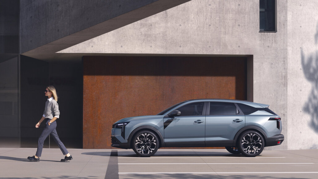 Ein hellblauer Crossover-SUV ist in der Seitenansicht vor einer modernen Beton- und Rostwand geparkten, während eine Frau im Vordergrund vorbeigeht.