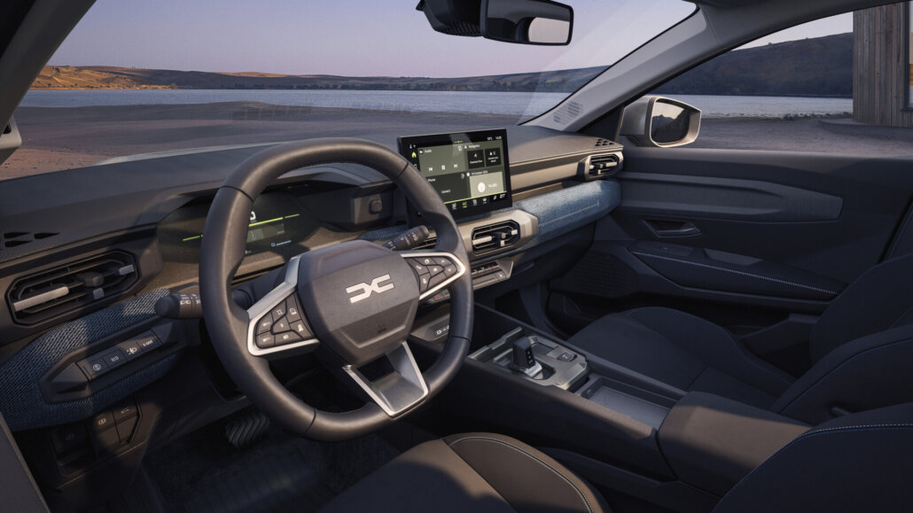 Der Innenraum eines dunkelgrauen Dacia Striker mit schwarzem Lenkrad und Infotainment-Bildschirm, fotografiert aus der Perspektive vom Fahrersitz, mit Blick auf eine Landschaft mit Gewässer und Hügeln.