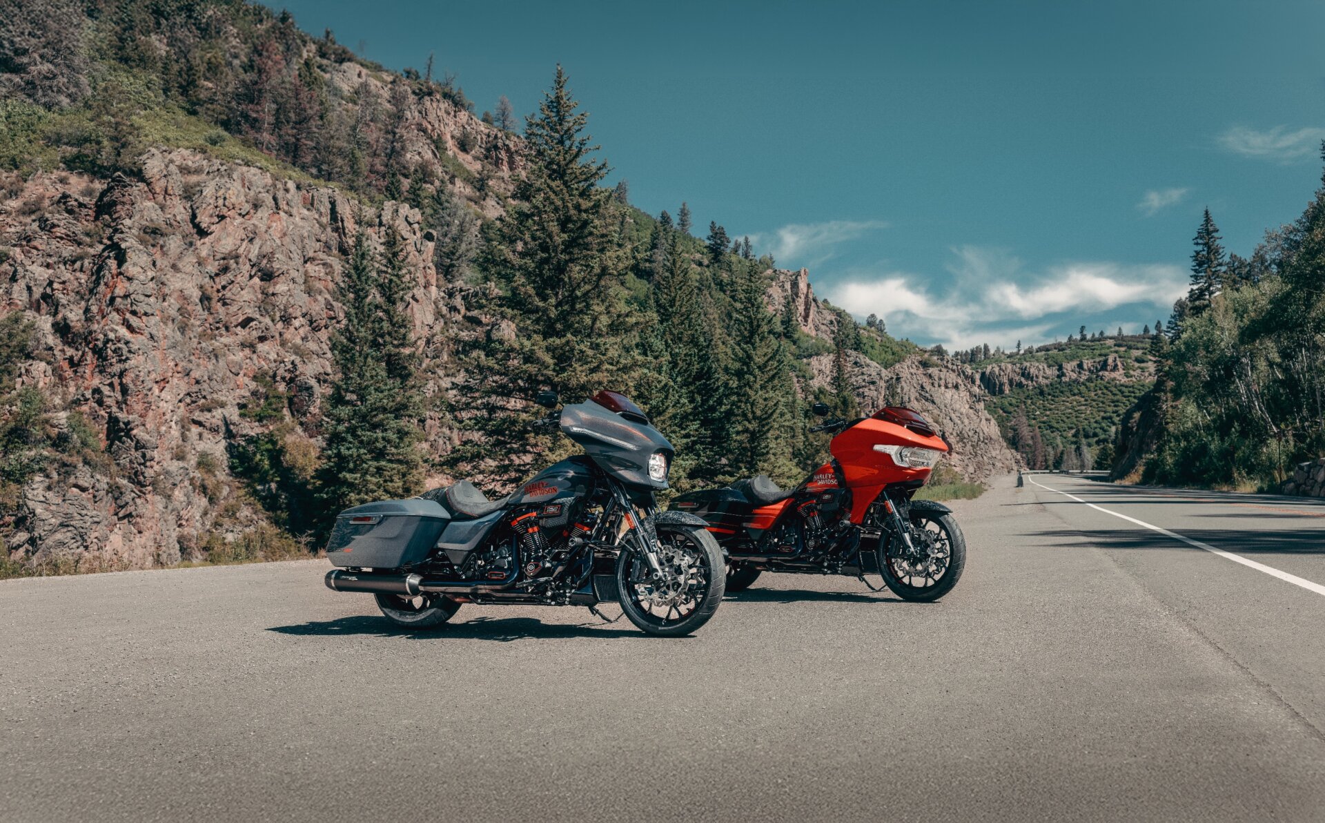 Zwei Harley-Davidson Touring-Motorräder mit Verkleidung und Koffern, eines dunkelgrau, eines orange, auf einer Straße vor Felslandschaft