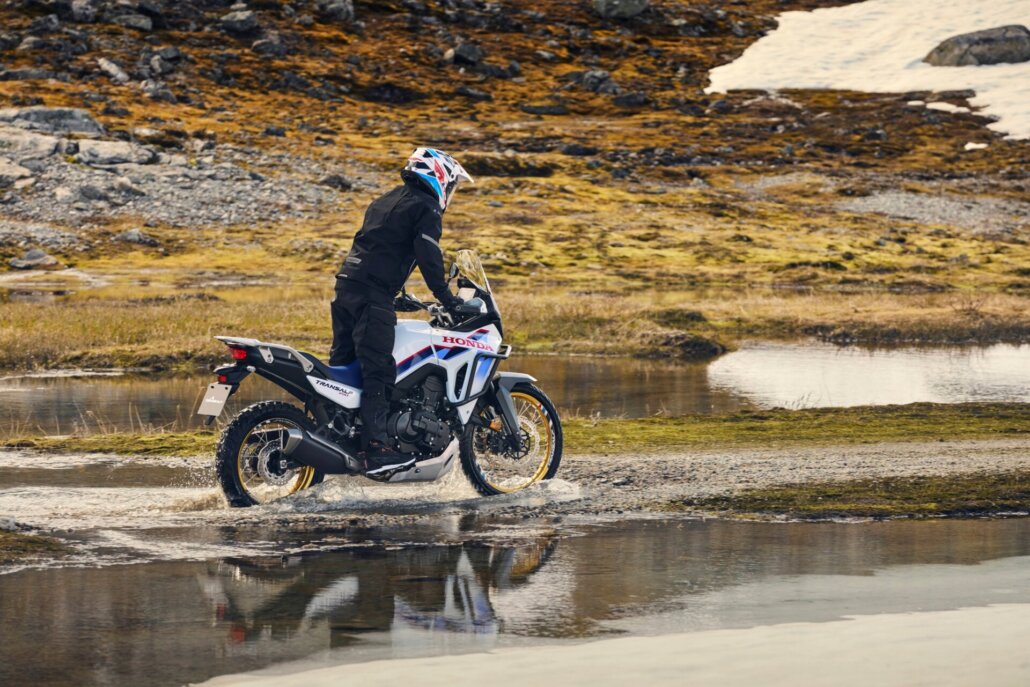 Dreiviertelansicht hinten rechts von einer weiß-blau-roten Honda Transalp 750, die durch eine flache Wasserstelle in einer kargen Natur- und Schneelandschaft fährt.