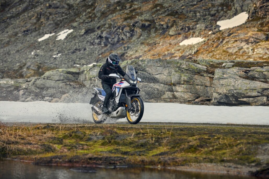 Dreiviertelansicht vorne rechts einer weiß-blau-roten Honda Transalp Enduro auf einer nassen Straße vor einer felsigen Berglandschaft mit Schneeflecken.