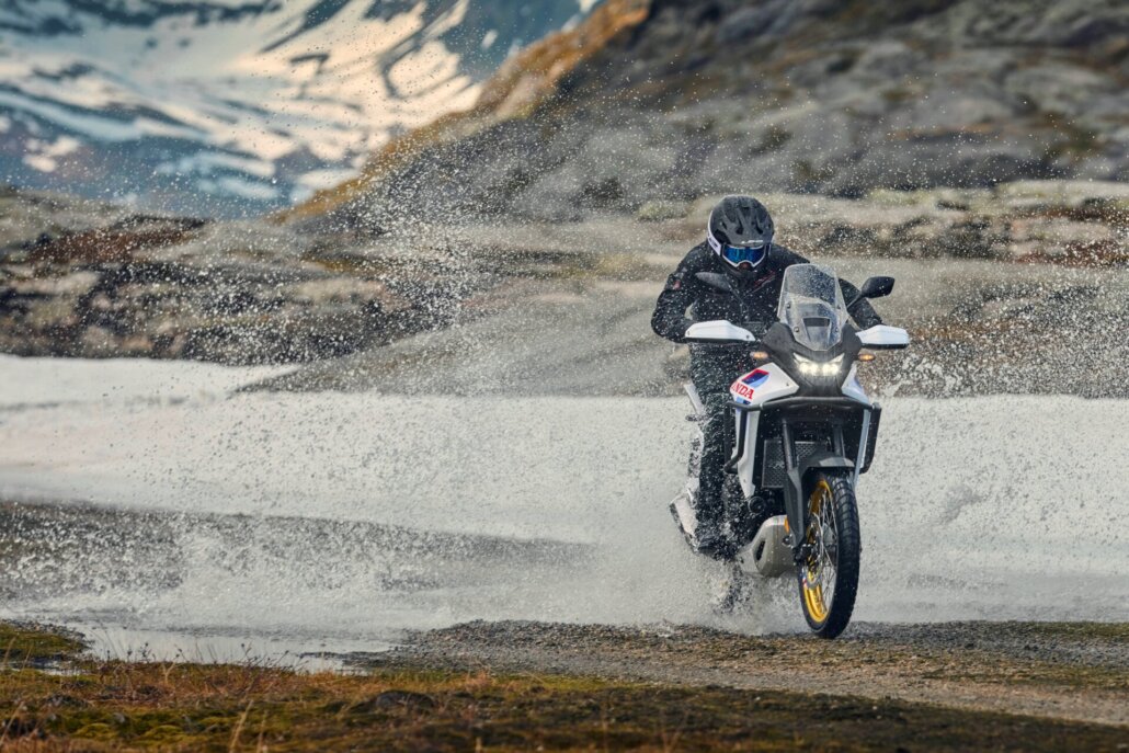 Ein weiß-schwarzes Honda-Motorrad in Frontalansicht fährt durch eine Pfütze in einer felsigen Berglandschaft mit Schneeresten und wirbelt dabei viel Wasser auf.
