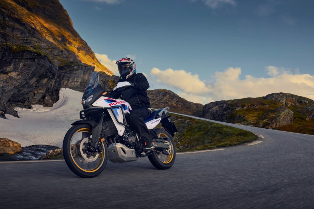 Dreiviertelansicht vorne links einer weiß-schwarz-blauen Honda Transalp 750 mit goldenen Felgen und fahrender Person in Schutzkleidung auf einer bergigen Passstraße mit Restschnee.