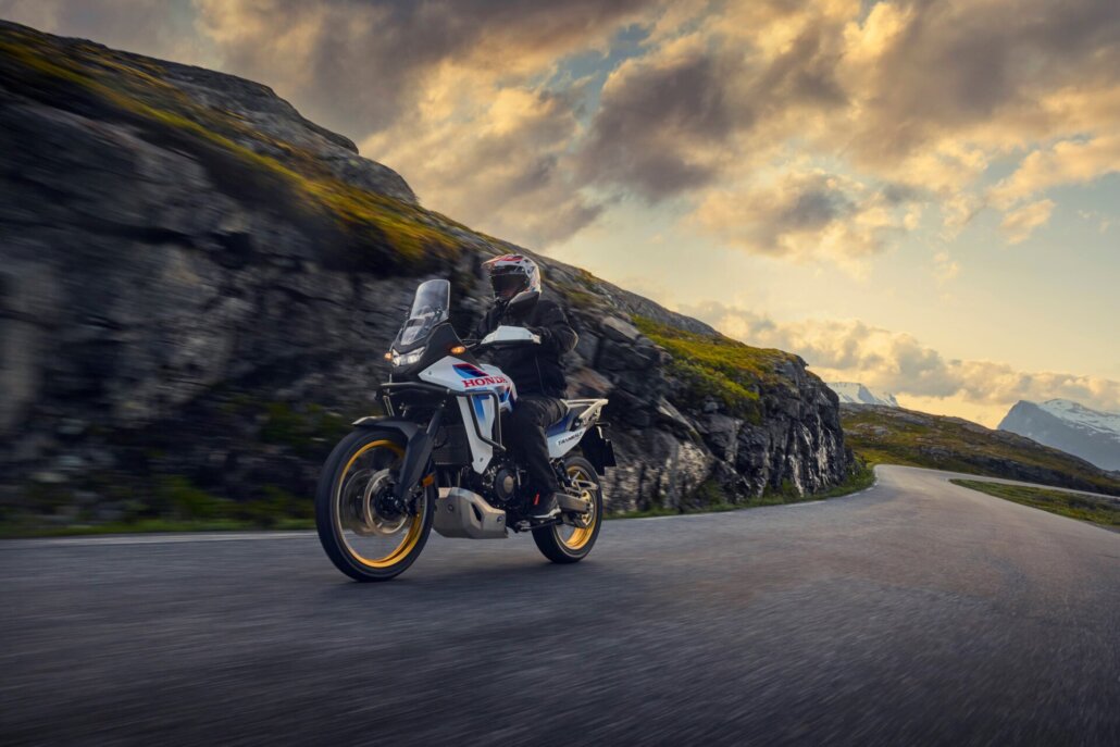 Ein weißes, blaues und rotes Honda Transalp Motorrad in Dreiviertelansicht vorne links fährt auf einer asphaltierten Küsten- oder Gebirgsstraße vorbei an felsigen Hängen bei bewölktem Himmel und Abendlicht.