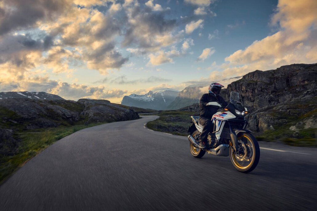 Eine weiß-blau-rote Honda-Reiseenduro mit goldenen Felgen in Dreiviertelansicht vorne rechts fährt auf einer gewundenen Asphaltstraße in einer gebirgigen Landschaft unter bewölktem Himmel.