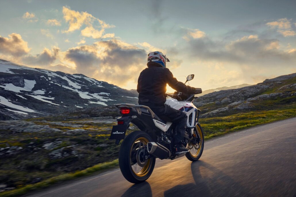 Eine weiß-schwarze Reiseenduro mit goldenen Felgen und dem Schriftzug „Transalp 750“ fährt in der Dreiviertelansicht von hinten auf einer Asphaltstraße durch eine bergige Landschaft mit Schneefeldern im Abendlicht.