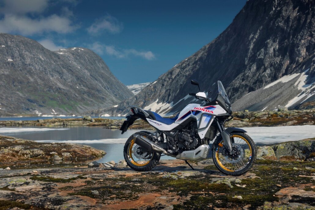 Eine weiß-blau-rote Honda Transalp 750 in Dreiviertelansicht vorne rechts auf einem felsigen Ufer vor einer alpinen Bergkulisse mit See und Schneeflecken.