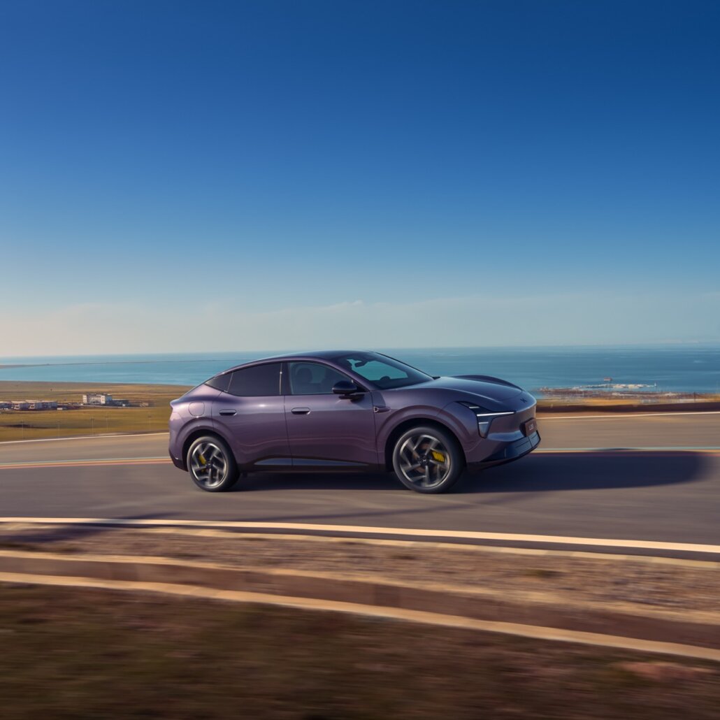 Ein lilafarbener Crossover-SUV in Dreiviertelansicht von vorne rechts fährt auf einer Straße an einer Küstenlandschaft unter klarem blauem Himmel.