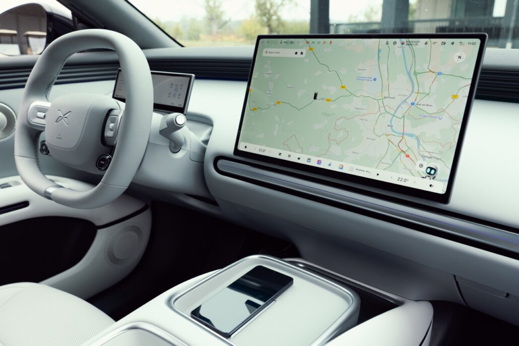 Das Foto zeigt den hellen Innenraum eines Autos in einer Frontalperspektive auf das Armaturenbrett, mit einem großen zentralen Touchscreen, der eine Straßenkarte anzeigt, einem hellgrauen Lenkrad und einer Ablagefläche mit einem Smartphone in der Mittelkonsole.
