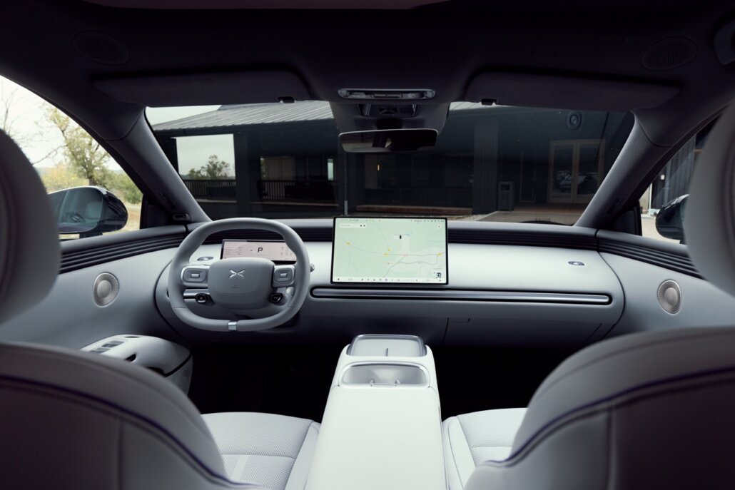 Innenraum eines Autos mit hellem Interieur, großem Zentralbildschirm und Lenkrad in Frontalperspektive, gesehen durch die Frontsitze auf eine dunkle Gebäudeumgebung.